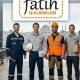 Four factory workers stand together in a workshop under a Fatih logo banner, one in a safety vest holding a helmet and a clipboard, others with boxes and tools nearby. -