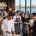 Restaurant scene with waitstaff wearing Fatih workwear aprons and chef coats; logo 'fatih' in top-left, panoramic window view of city by the water in the background.