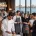 Restaurant scene with waitstaff wearing Fatih workwear aprons and chef coats; logo 'fatih' in top-left, panoramic window view of city by the water in the background. -