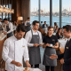 gastronomi ve turizm aşçı servis kıyafetleri - Fatih iş elbiseleri istanbul Restaurant scene with waitstaff wearing Fatih workwear aprons and chef coats; logo 'fatih' in top-left, panoramic window view of city by the water in the background. -
